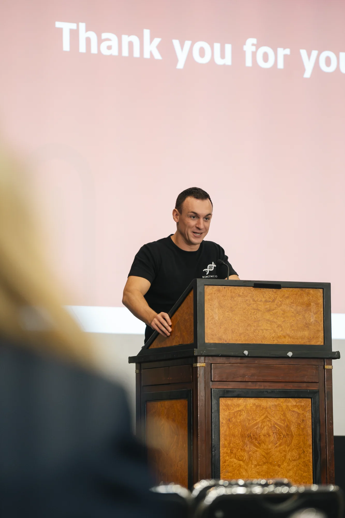 A conference presenter at the lectern in a DoNotAge.org shirt